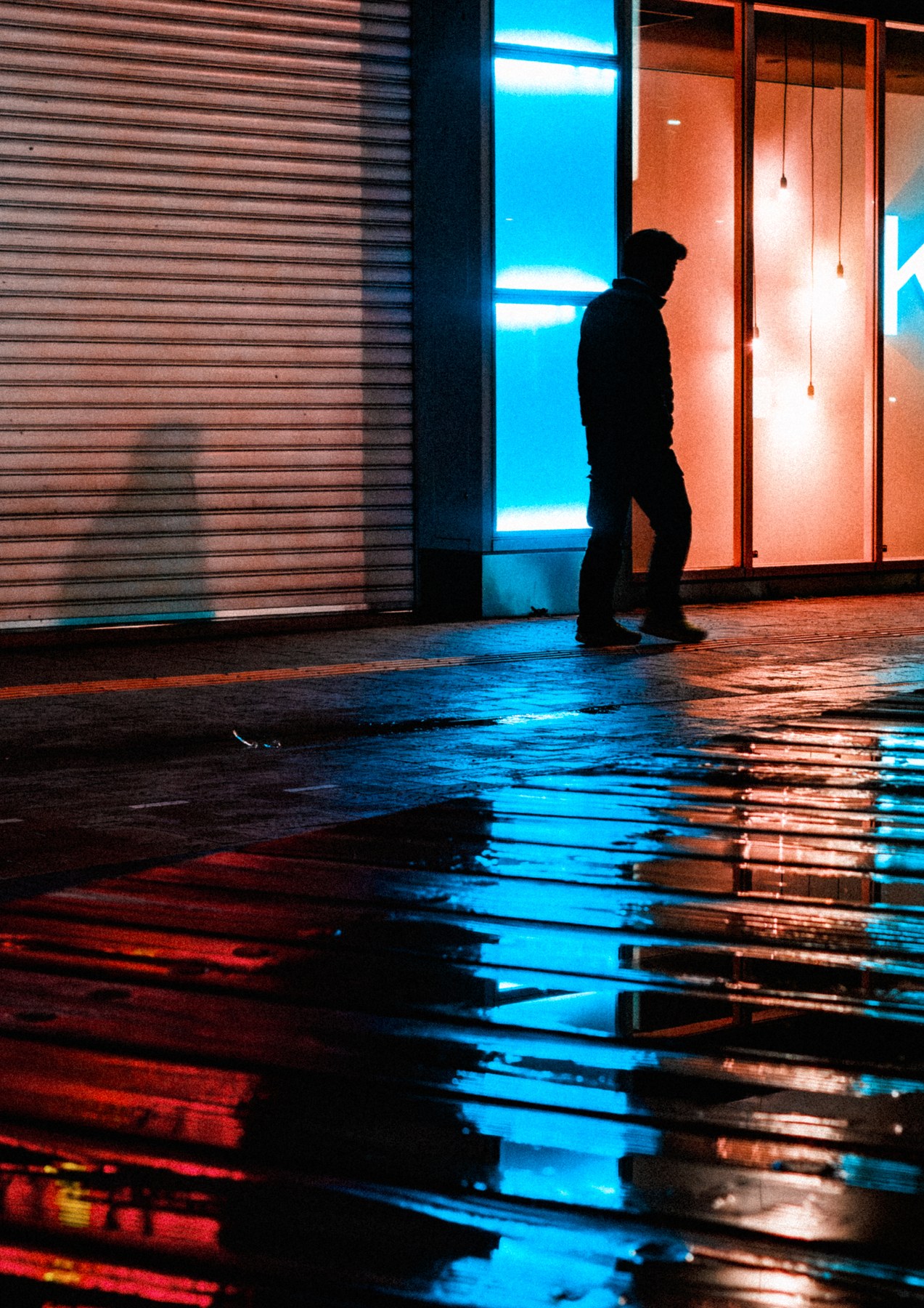 雨に濡れた路面に映る青と橙の光の間を歩く人影。