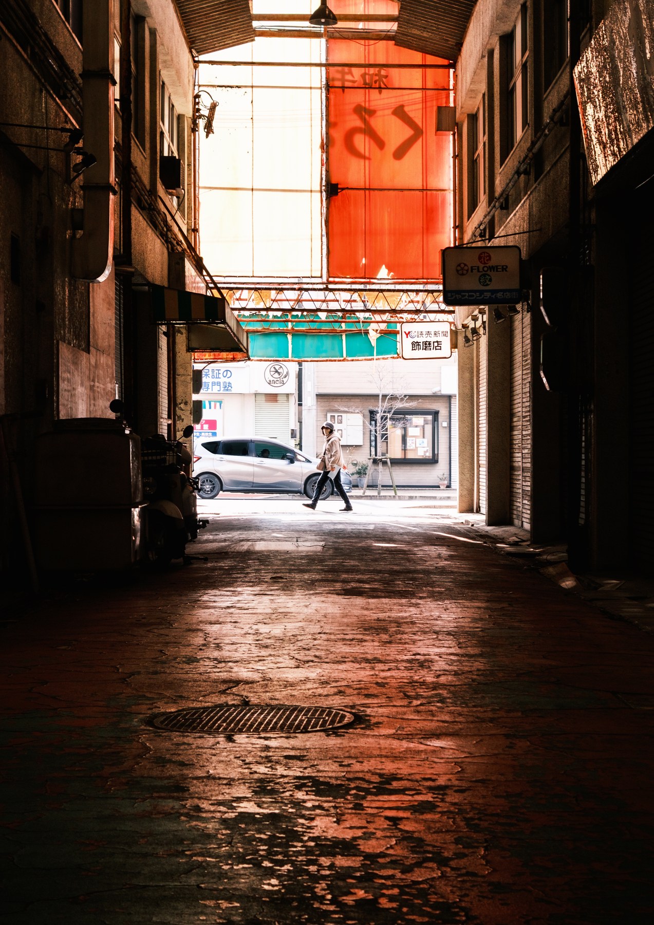 A person walking through a dim covered passage toward a bright street beyond.
